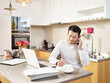 telecommuting young asian business person sitting at kitchen counter at home talking on cellphone
