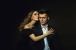 Beautiful young stylish couple, man and woman, in black evening dresses in each other's arms posing in the studio on a black background.