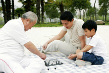 A Boy Playing Chess With His Grandfather On The Lakeside While His Father Is Watching