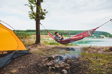 Wall Mural - man laying on hammock at lake beach near camp fire