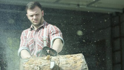 Wall Mural - Portrait of caucasian young man worker sawing a log with a chainsaw. Video. Male carpenter sawing a part of tree trunk with wooden shavings flying into the sides.