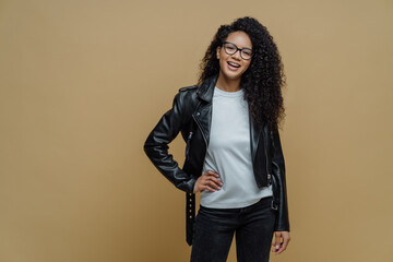 Wall Mural - Fashionable woman with glad expression, keeps hand on hip, smiles cheerfully, wears optical glasses, white t shirt, black leather jacket and jeans, ready for walk, poses against beige background