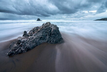 Long Exposure Of Water Flowing Between A Rock On The Beach With Stunning Clouds In The Background