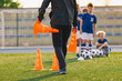 © matimix - School coach preparing field for soccer training. Kids waiting in line in the background. Trainer holding training cones. Sporst school stadium in the background