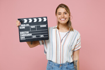 Wall Mural - Smiling young blonde woman girl in casual striped shirt posing isolated on pink background studio portrait. People lifestyle concept. Mock up copy space. Hold classic black film making clapperboard.