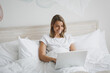 © ViDi Studio - Smiling young woman girl lying in bed with white sheet pillow blanket spending time in bedroom at home. Rest relax good mood lifestyle concept. Mock up copy space. Working on laptop pc computer.