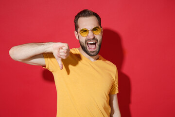 Crazy young bearded man guy in casual yellow t-shirt glasses posing isolated on red wall background studio portrait. People sincere emotions lifestyle concept. Mock up copy space. Showing thumb down.