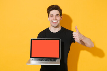 Funny young man guy 20s in casual black t-shirt posing isolated on yellow background. People lifestyle concept. Mock up copy space. Hold laptop pc computer with blank empty screen, showing thumb up.