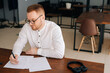 © dikushin - Talented young composer wearing stylish eyeglasses writing musical notes with pen on paper sheet music sitting at the wooden desk in modern room in office or home, headphones on table