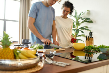 Young man cutting vegetables while woman holding pepper and watching him. Vegetarians preparing healthy meal in the kitchen together. Vegetarianism, healthy food concept