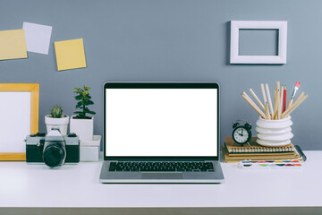 Front view of laptop with blank white screen, film retro camera, paints and other offive supplies on the white office desk