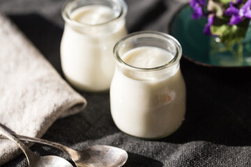 Two glass jars with natural homemade yogurt, spoons, little vase with violets on black fabric on white wooden table.