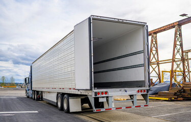 Big rig blue semi truck with open door semi trailer standing at warehouse parking lot at industrial area waiting for the load for the next delivery