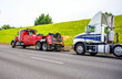 Red big rig semi towing truck prepare to tow broken white big rig semi tractor standing out of service on the road side