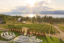 Wedding In The Winery. Beautiful Romantic Set Up. Decorated Chairs And Wedding Arbor In Front Of Vineyards By Okanagan Lake In Kelowna. The Sun Shining Through Clouds During Sunset In The Background.