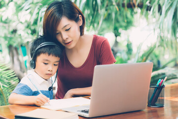 Wall Mural - Young asian mother and son using laptop computer for study and learning together at home, boy writing on notebook for homework and wearing headphone, teacher or mom support child, education concept.