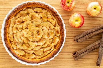 Wall Mural - Homemade baked French apple tart, an open faced apple pie, in a baking white ceramic dish aside Gala red apples and cinnamon sticks, all on an oak wood background. Flat lay, top view.