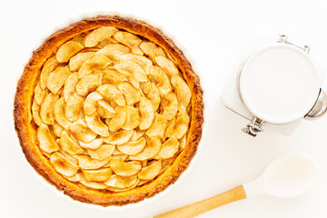 Wall Mural - Homemade baked French apple tart, an open faced apple pie, in a baking white ceramic dish aside a white cooking spoon and a white jar on a white background. Flat lay, top view with copy space.