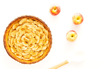 Wall Mural - Homemade baked French apple tart, an open faced apple pie, in a baking white ceramic dish aside Gala apples and a white cooking spoon on a white background. Flat lay, top view.