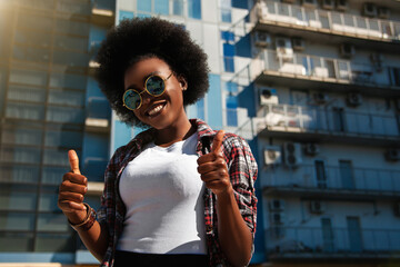 Image of a happy optimistic emotional young american african student posing over city background dressed casual and wearing sunglasses, showing thumbs up gesture. Lifestyle.