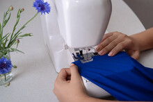 Girl Sews A Dress Of Blue Chiffon On A Sewing Machine, Near A Sewing Machine There Is A Cornflowers In A Vase. Woman Using The Sewing Machine To Sew