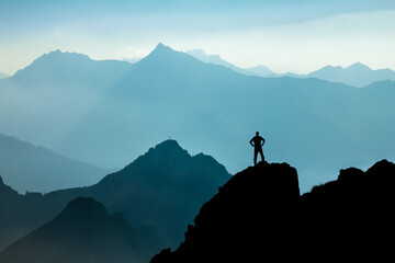 Aufkleber - Man reaching summit after climbing and hiking enjoying freedom and looking towards mountains silhouettes panorama during sunrise.