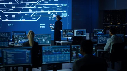Female Project Leader Using Digital Tablet Computer and Looking at Big Screen Display Showing Infrastructure Infographics and Data.Telecommunications Control Monitoring Room with People Working
