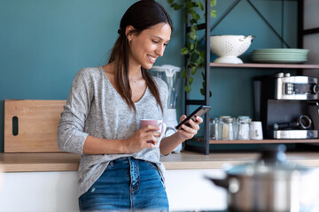 Smiling young woman using her mobile phone while drinking a cup of coffee in the kitchen at home.