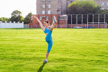 Overall Plan. Beautiful Brunette Girl Athlete, Dressed In A Top And Tight Leggings, Doing Sports Exercises On A City Field. Raising Leg Up, Casting A Shadow On The Grass. Stretching, Training Her Body