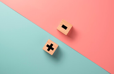 Black of plus and minus sign in opposite side of wooden cube on blue and pink background.