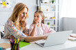 © LIGHTFIELD STUDIOS - Selective focus of smiling woman using laptop near kid holding pen during online education