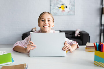 Wall Mural - Selective focus of smiling kid holding laptop and looking at camera near stationery