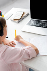 Wall Mural - Selective focus of kid in headset writing on notebook during electronic education at home