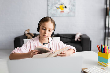 Wall Mural - Selective focus of child in headset reading book near laptop on table