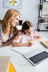 Wall Mural - Selective focus of smiling woman embracing kid writing on notebook near laptop on table