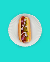 Flat Lay Hot Dog In Bun Still Life. Fankfurter In Bun With Relish And Onions On A White Paper Plate On Teal Background With Copy Space.