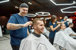 © Svitlana - Cheerful professional tattooed barber making haircut for a young and happy handsome man sitting in a barbershop chair. Focus on a smiling guy