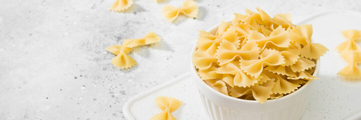 raw farfalle pasta in a white bowl on a light gray table. pasta farfalle close-up. banner