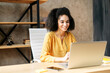 Mixed-race girl with an afro hairstyle uses hands free headset for online communication with a customers or coworkers indoors, she looks at laptop screen and smiles