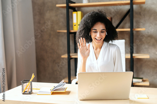 Cheerful biracial young woman uses laptop for video call. A glad girl sits in modern office and waving hello into webcam. Front view. Video meeting concept