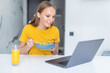 © F8  \ Suport Ukraine - Beautiful young woman eat breakfast leaning on the kitchen table with laptop.