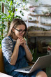 © DimaBerlin - Woman gardener in glasses wear blue jeans overalls, sitting on chair in greenhouse, using laptop after work, communicates on internet with customer surrounded by plants. Home gardening, freelance.