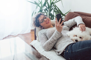 Young woman using mobile phone. Young woman laying on sofa with her dog and using mobile phone.