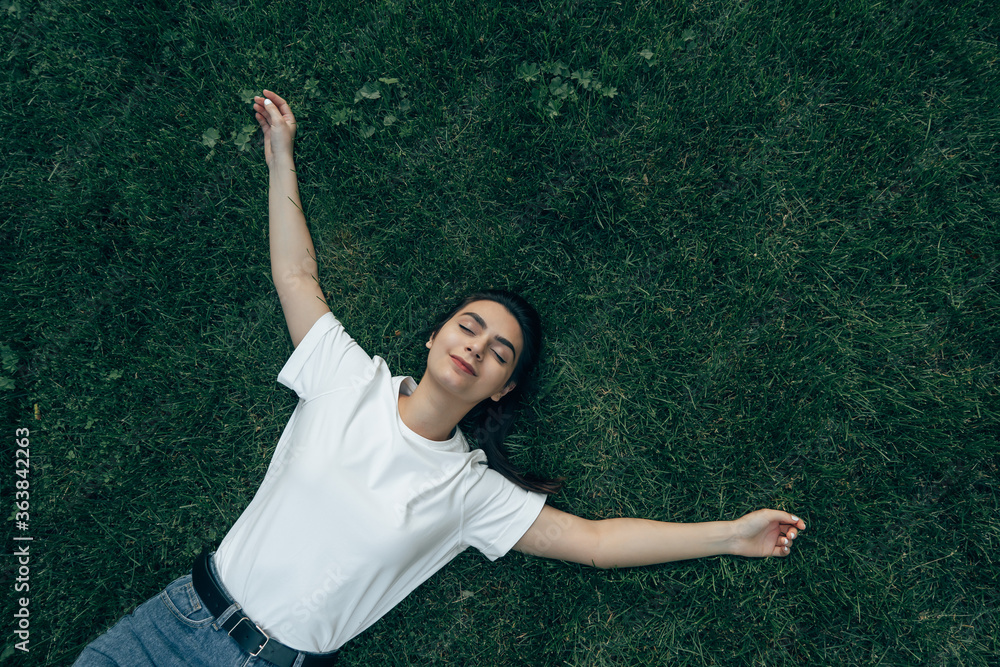 Caucasian girl 20-25 years old lies on the green grass on a summer day smiling, arms outstretched, hugging everyone. View from above. View from the corner. The concept of a happy life, vacation