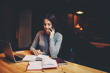 Young businesswoman talking on mobile phone with friend and making accounting reports sitting in own coffee shop at wooden table with laptop computer device connected to wireless 4G internet