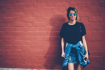 Attractive hipster girl 20 years old casual dressed looking at camera while standing near wall and holding modern mobile phone in hand, concept of trendy urban fashion woman and street denim style