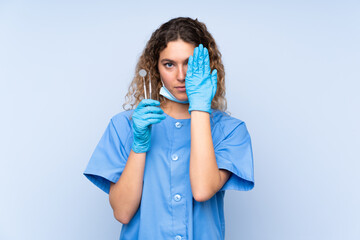 Wall Mural - Young blonde woman dentist holding tools isolated on blue background covering a eye by hand