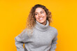 Young blonde woman with curly hair wearing a turtleneck sweater isolated on yellow background posing with arms at hip and smiling