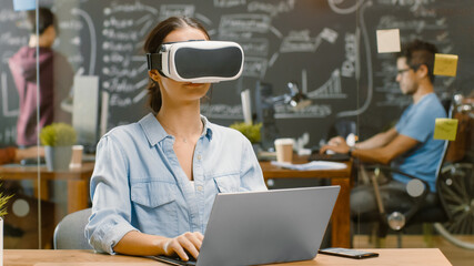 Wall Mural - young female creative developer wearing virtual reality headset works on a laptop, she develops new 