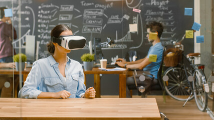 Wall Mural - young female creative developer wearing virtual reality headset works on a laptop, she develops new 